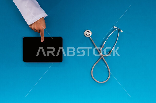 Marketing for modern gadgets, vertical overhead portrait of a Saudi Arabian Gulf man conducting research and studies on a tablet and next to a stethoscope, blank black screen display, online health consultation request, concept of medicine and healthcare, medical services and devices, blue background