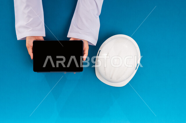 Using modern technology in engineering work, Displaying a blank black screen, Buying and selling supplies online, Training via the Internet, Vertical top portrait of a Saudi Arabian Gulf man holding a tablet and next to him a white protective helmet, Working in the engineering sector, Blue background