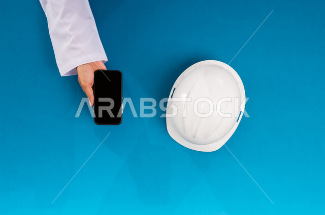 بيع وشراء المستلزمات أونلاين، التدريب من خلال الانترنت، بورتريه علوي عامودي ليد رجل عربي سعودي خليجي يمسك جهاز الجوال وبجانبه خوذة الحماية البيضاء، العمل في القطاع الهندسي، استخدام الأجهزة التقنية الحديثة بالأعمال الهندسية، عرض شاشة سوداء فارغة، خلفية زرقاء