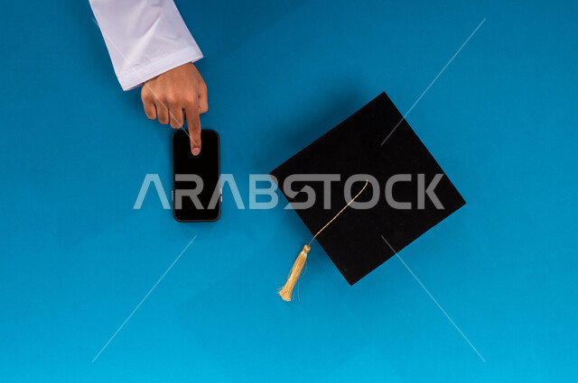 الاستفادة من التكنولوجيا الحديثة لمشروع التخرج، البيع والشراء عبر الانترنت، استخدام تطبيقات التسوق عن بعد، طلب مستلزمات التخرج أونلاين، عرض شاشة سوداء فارغة، بورتريه علوي عامودي ليد رجل عربي سعودي خليجي يستخدم الهاتف المحمول وبجانبه قبعة تخرج، خلفية زرقاء