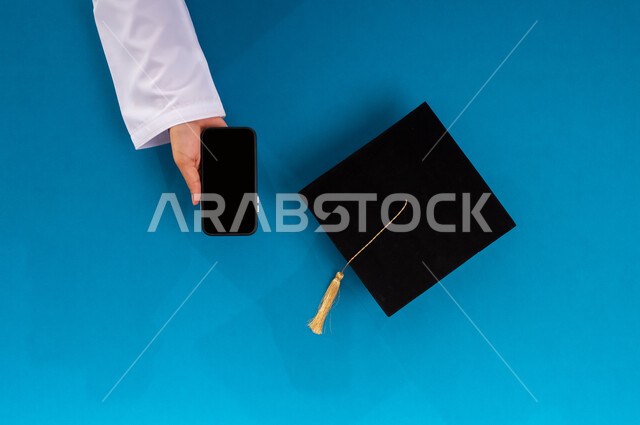 البيع والشراء عبر الانترنت، استخدام تطبيقات التسوق عن بعد، طلب مستلزمات التخرج أونلاين، عرض شاشة سوداء فارغة، بورتريه علوي عامودي ليد رجل عربي سعودي خليجي يمسك الهاتف المحمول وبجانبه قبعة تخرج، الاستفادة من التكنولوجيا الحديثة لمشروع التخرج، خلفية زرقاء