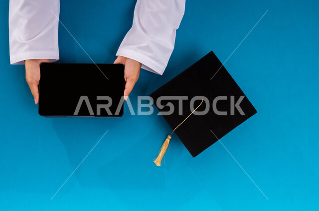 استخدام تطبيقات التسوق عن بعد، طلب مستلزمات التخرج أونلاين، عرض شاشة سوداء فارغة، بورتريه علوي عامودي ليد رجل عربي سعودي خليجي يمسك التابليت وبجانبه قبعة تخرج، الاستفادة من التكنولوجيا الحديثة لمشروع التخرج، البيع والشراء عبر الانترنت، خلفية زرقاء