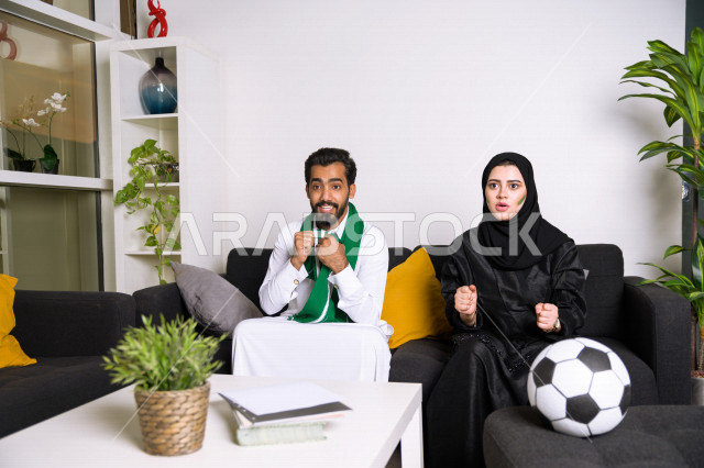 زوجان عربيان سعوديان خليجيان يشاهدان مباراة للدوري السعودي ، يشجعان فريق الأهلي و السعودية , ايماءات وجه تدل على الحماس والفرحة بالفوز , أندية وتشجيع .