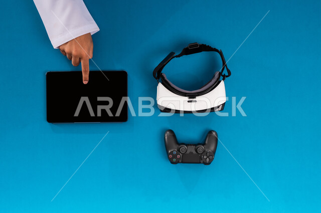 Using modern technical devices, simulating the virtual world, technological and technical development in the Kingdom, enjoying video games, displaying a blank black screen, vertical upper portrait of the hands of a Saudi Arabian Gulf man working on a tablet and next to him are VR glasses and a joystick, blue background