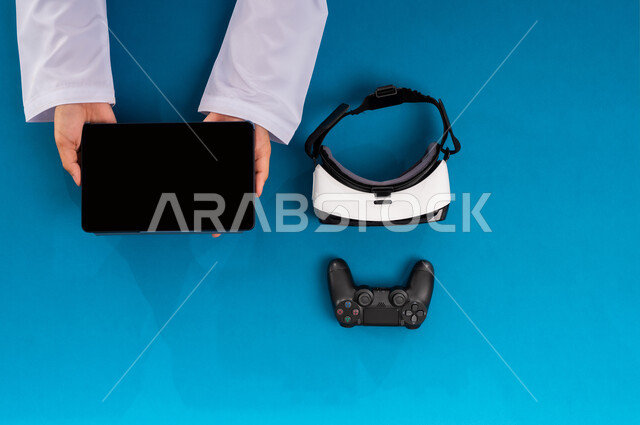 Enjoying video games, showing a blank black screen, vertical overhead portrait of a Saudi Arabian Gulf man holding a tablet and next to him VR glasses and controller, using modern technical devices, simulating the virtual world, technological and technical development in the Kingdom, blue background