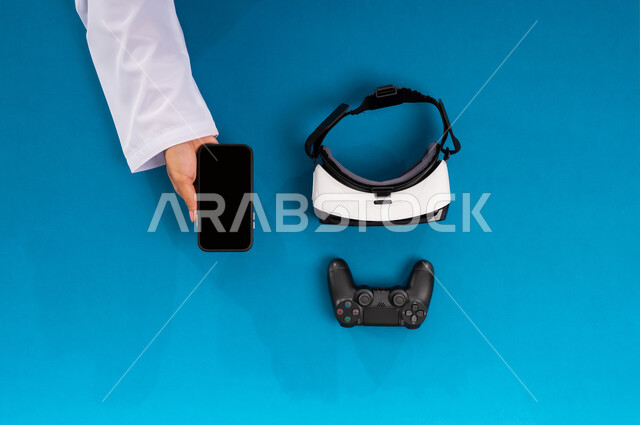 Technological and technical development in the Kingdom, enjoying video games, displaying a blank black screen, vertical overhead portrait of the hands of a Saudi Arabian Gulf man holding a mobile device and next to him VR glasses and a joystick, using modern technical devices, simulating the virtual world, blue background