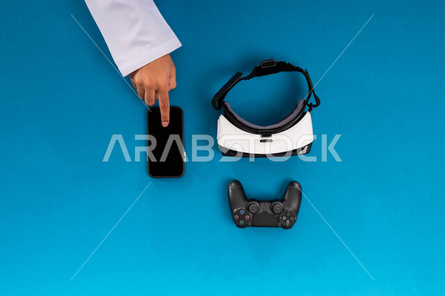 محاكاة العالم الافتراضي، التطور التكنولوجي والتقني في المملكة، الاستمتاع في العاب الفيديو، عرض شاشة سوداء فارغة، بورتريه علوي عامودي ليدين رجل عربي خليجي سعودي يعمل على الجوال وبجانبه نظارة الفي آر وقبضة التحكم، استخدام الأجهزة التقنية الحديثة, خلفية زرقاء