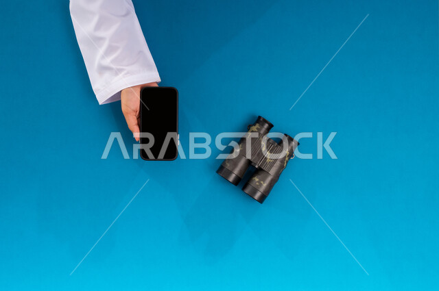 التخطيط المستقبلي للأهداف والطموحات، مفهوم النظرة المستقبلية، عرض شاشة سوداء فارغة، بورتريه علوي عامودي ليدين رجل عربي خليجي سعودي يحمل جهاز الجوال وبجانبه المنظار ثنائي العينين، استعمال المنظار للكشف عن الاشياء البعيدة, حب الاستكشاف والمغامرة،خلفية زرقاء