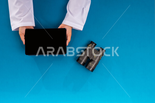مفهوم النظرة المستقبلية، عرض شاشة سوداء فارغة، بورتريه علوي عامودي ليدين رجل عربي خليجي سعودي يحمل جهاز التابليت وبجانبه المنظار ثنائي العينين، استعمال المنظار للكشف عن الاشياء البعيدة, حب الاستكشاف والمغامرة، التخطيط المستقبلي للأهداف والطموحات، خلفية زرقاء
