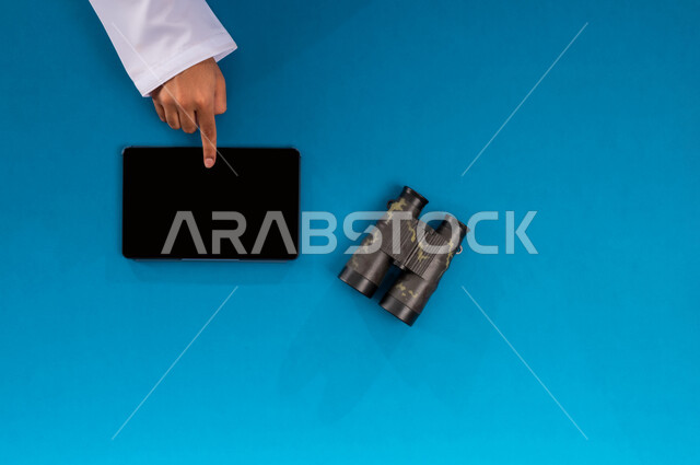 استعمال المنظار للكشف عن الاشياء البعيدة, حب الاستكشاف والمغامرة، التخطيط المستقبلي للأهداف والطموحات، مفهوم النظرة المستقبلية، عرض شاشة سوداء فارغة، بورتريه علوي عامودي ليدين رجل عربي خليجي سعودي يستخدم جهاز التابليت وبجانبه المنظار ثنائي العينين، خلفية زرقاء
