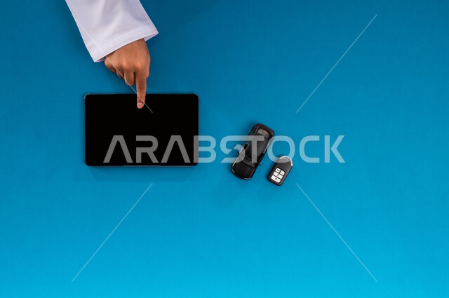 عرض شاشة سوداء فارغة، البيع والشراء من الانترنت، بورتريه علوي عامودي ليد رجل عربي خليجي سعودي يعمل على جهاز التابليت وبجانبه مجسم صغير لسيارة ومفتاح، الحصول على السيارات الحديثة اونلاين، استخدام التكنولوجيا في طلب المستلزمات، خلفية زرقاء