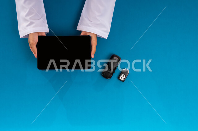 استخدام التكنولوجيا في طلب المستلزمات، عرض شاشة سوداء فارغة، البيع والشراء من الانترنت، بورتريه علوي عامودي ليد رجل عربي خليجي سعودي يمسك جهاز التابليت وبجانبه مجسم صغير لسيارة ومفتاح، الحصول على السيارات الحديثة اونلاين، خلفية زرقاء