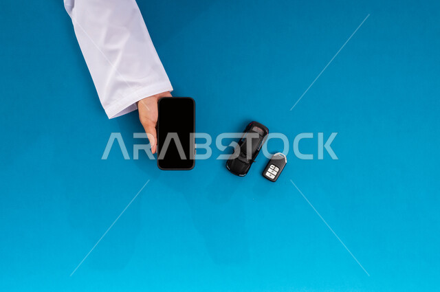 الحصول على السيارات الحديثة اونلاين، استخدام التكنولوجيا في طلب المستلزمات، عرض شاشة سوداء فارغة، البيع والشراء من الانترنت، بورتريه علوي عامودي ليد رجل عربي خليجي سعودي يمسك جهاز الهاتف المحمول وبجانبه مجسم صغير لسيارة ومفتاح، خلفية زرقاء