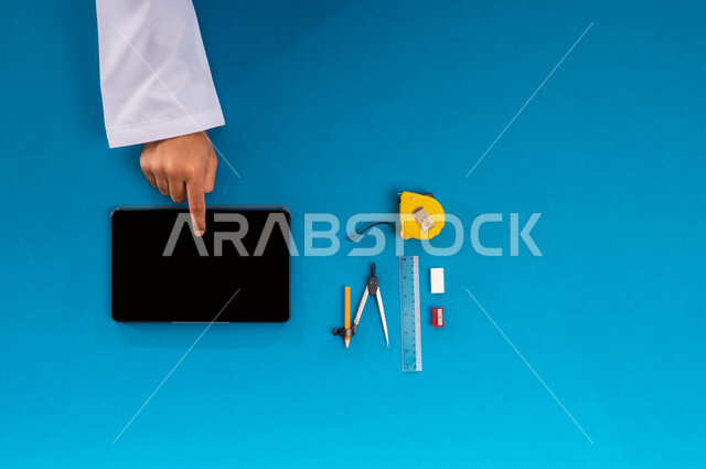 دمج التقنيات الحديثة بالعمل، استخدام المواقع الالكترونية للبحث عن مستلزمات البناء، بورتريه علوي عامودي ليدي رجل عربي خليجي سعودي يعمل على جهاز التابليت وبجانبه الأدوات الهندسية، البيع والشراء والتسوق عبر الانترنت، عرض شاشة سوداء فارغة، خلفية زرقاء
