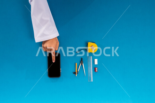 استخدام المواقع الالكترونية للبحث عن مستلزمات البناء، بورتريه علوي عامودي ليدي رجل عربي خليجي سعودي يستخدم جهاز الهاتف المحمول وبجانبه الأدوات الهندسية، التسوق والبيع والشراء عبر الانترنت، عرض شاشة سوداء فارغة، دمج التقنيات الحديثة بالعمل، خلفية زرقاء