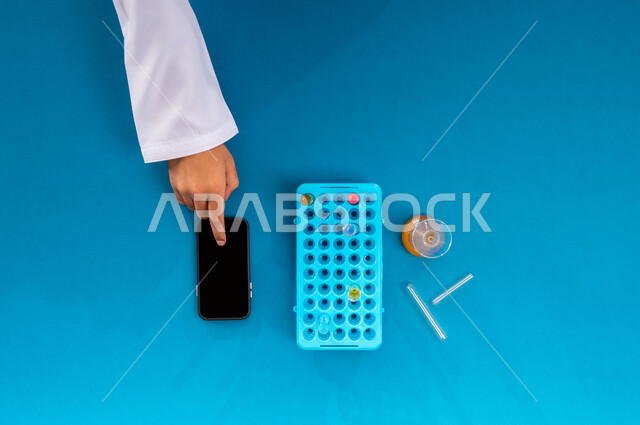 Scientific research and development in the field of chemistry, blank black screen display, vertical overhead portrait of a Saudi Arabian Gulf man working on a mobile device with test tubes next to him, recording laboratory test results on the mobile, conducting scientific experiments and research through biotechnology, blue background