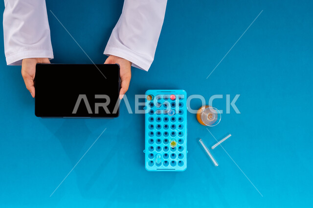 Recording laboratory test results on tablet, conducting scientific experiments and research through biotechnology, scientific research and development in the field of chemistry, displaying a blank black screen, vertical top portrait of a Saudi Arabian Gulf man's hand holding an iPad and test tubes next to him, blue background