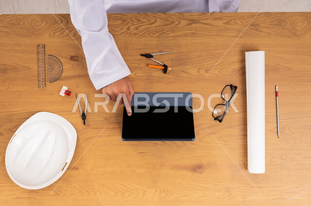Shopping through websites, A vertical top view of the hands of a Saudi Arabian Gulf man using a tablet with a protective helmet and engineering tools next to him, Displaying a blank black screen, Obtaining the necessary supplies online, Concept of engineering and architectural construction, Integrating modern technologies into work