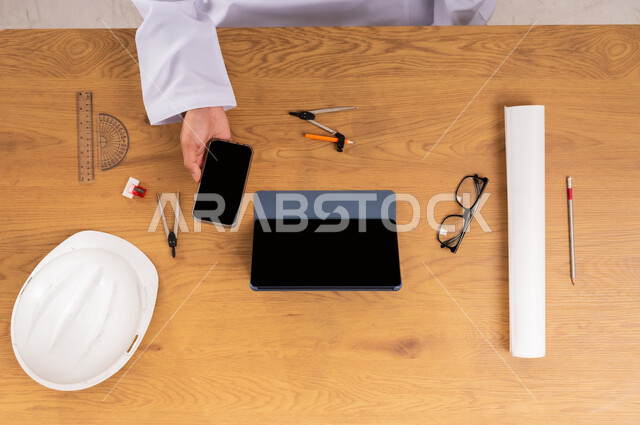 مفهوم الهندسة والإنشاءات المعمارية، التسوق عبر المواقع الالكترونية، صورة علوية عامودية ليدين رجل عربي خليجي سعودي يستخدم جهاز الهاتف المحمول والتابليت  وبجانبه خوذة الحماية والأدوات الهندسية، عرض شاشة سوداء فارغة، الحصول على المستلزمات اللازمة اونلاين