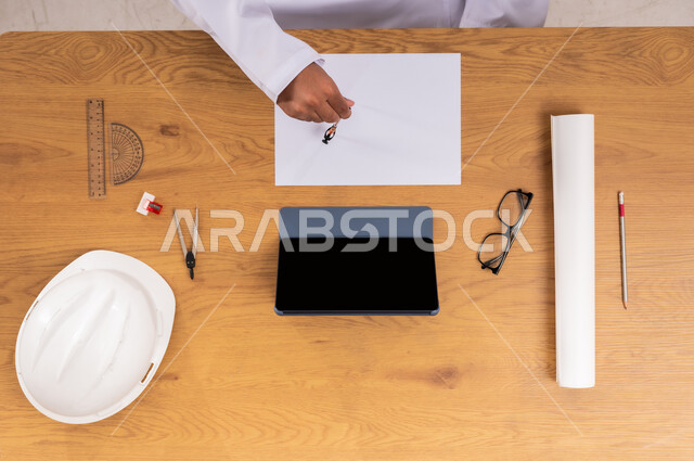 Reviewing and implementing construction projects, Obtaining building supplies through websites, Displaying a blank black screen on a tablet, A vertical overhead image of the hands of a Saudi Arabian Gulf man working on drawing an engineering plan with a protective helmet and engineering tools next to him, Building engineering using modern methods