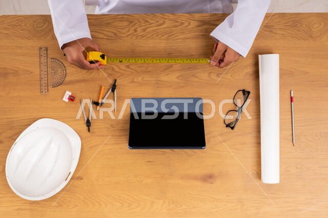 أخذ القياسات المناسبة بالمتر, استخدام أدوات و معدات الهندسة, دمج التقنيات الحديثة بالأعمال الهندسية، صورة علوية عامودية ليدين مهندس عربي خليجي سعودي يمسك الشريط المتري بيديه, استعمال التكنولوجيا في الحياة العملية، عرض شاشة سوداء فارغة على جهاز التابليت