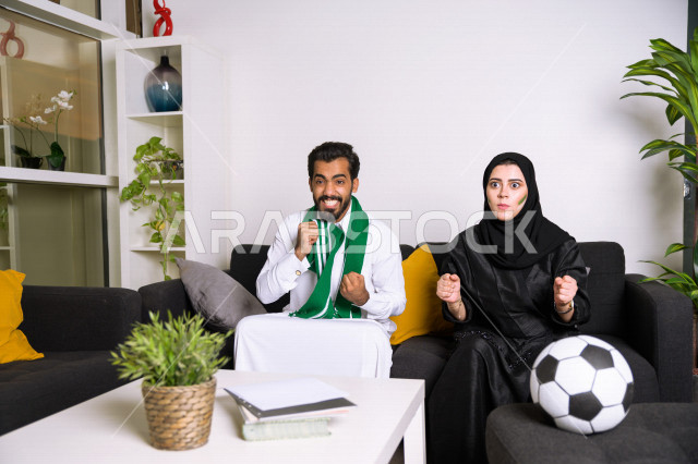 زوجان عربيان سعوديان خليجيان يشاهدان مباراة للدوري السعودي ، يشجعان فريق الأهلي و السعودية , ايماءات وجه تدل على الحماس والفرحة بالفوز , أندية وتشجيع .