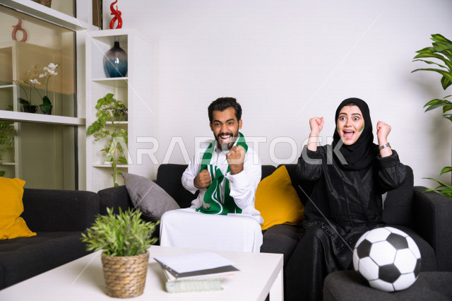 زوجان عربيان سعوديان خليجيان يشاهدان مباراة للدوري السعودي ، يشجعان فريق الأهلي و السعودية , ايماءات وجه تدل على الحماس والفرحة بالفوز , أندية وتشجيع .