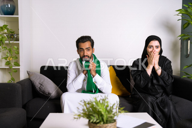 زوجان عربيان سعوديان خليجيان يشاهدان مباراة للدوري السعودي ، يشجعان فريق الأهلي و السعودية , ايماءات وجه تدل على الصدمة والخذلان بالخسارة , أندية وتشجيع .