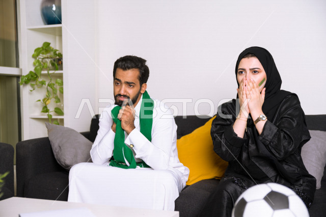 زوجان عربيان سعوديان خليجيان يشاهدان مباراة للدوري السعودي ، يشجعان فريق الأهلي و السعودية , ايماءات وجه تدل على الصدمة والخذلان بالخسارة , أندية وتشجيع .