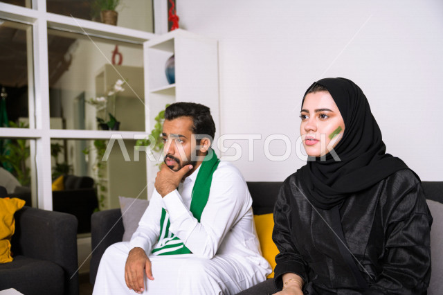 زوجان عربيان سعوديان خليجيان يشاهدان مباراة للدوري السعودي ، يشجعان فريق الأهلي و السعودية , ايماءات وجه تدل على الشغف والإنتماء للفريق , أندية وتشجيع .