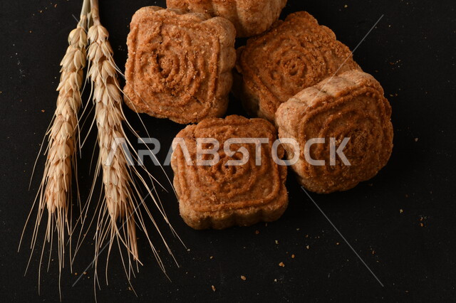 صورة لمجموعة من نباتات القمح، ومعمول محشو مربع الشكل من الحلويات الشعبية الهشة اللذيذة على خلفية سوداء ، صورة لكعك بعجوة وبعرف بإسم كعك العيد الشهي ، خلفية سوداء لحلويات شعبية طازجة