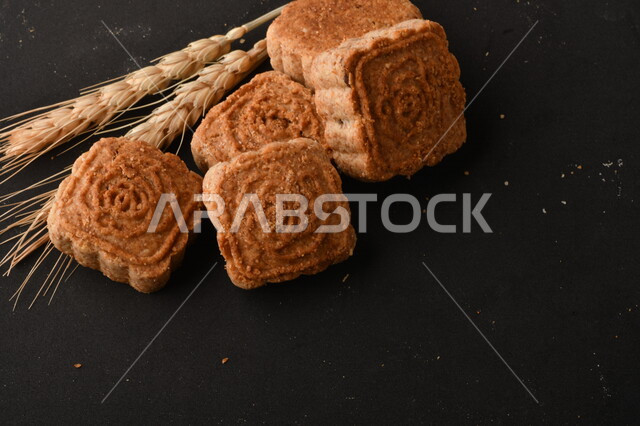 صورة لمجموعة من نباتات القمح، ومعمول محشو مربع الشكل من الحلويات الشعبية الهشة اللذيذة على خلفية سوداء ، صورة لكعك بعجوة وبعرف بإسم كعك العيد الشهي ، خلفية سوداء لحلويات شعبية طازجة