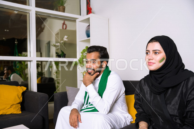 زوجان عربيان سعوديان خليجيان يشاهدان مباراة للدوري السعودي ، يشجعان فريق الأهلي و السعودية , ايماءات وجه تدل على الشغف والإنتماء للفريق , أندية وتشجيع .