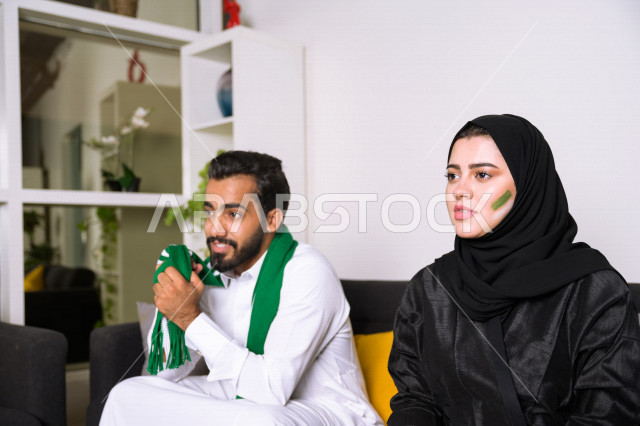 زوجان عربيان سعوديان خليجيان يشاهدان مباراة للدوري السعودي ، يشجعان فريق الأهلي و السعودية , ايماءات وجه تدل على الحماس والفرحة بالفوز , أندية وتشجيع .
