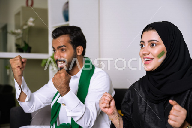 زوجان عربيان سعوديان خليجيان يشاهدان مباراة للدوري السعودي ، يشجعان فريق الأهلي و السعودية , ايماءات وجه تدل على الحماس والفرحة بالفوز , أندية وتشجيع .