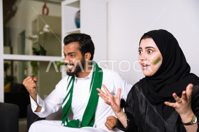 زوجان عربيان سعوديان خليجيان يشاهدان مباراة للدوري السعودي ، يشجعان فريق الأهلي و السعودية , ايماءات وجه تدل على الحماس والفرحة بالفوز , أندية وتشجيع .