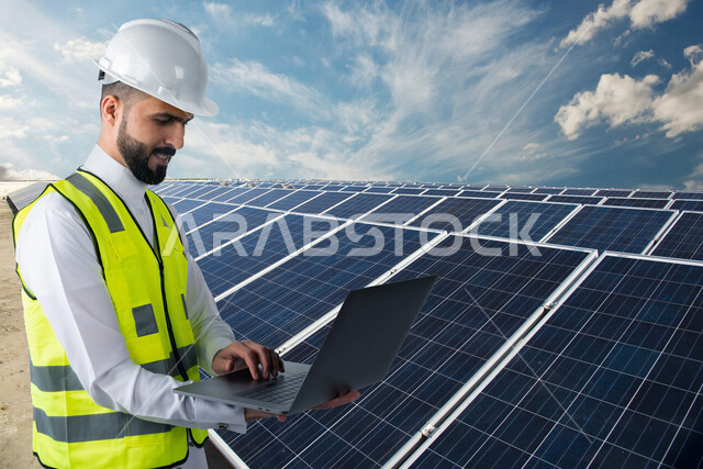صورة لمهندس كهربائي عربي خليجي سعودي يحمل حاسوب محمول في يده يقف بالقرب من ألواح للخلايا الشمسية ، استخدام الطاقة الشمسية لتوليد الكهرباء ، مفهوم استخدام الطاقة المتجددة ، مفهوم التنمية المستدامة بالطاقة البديلة