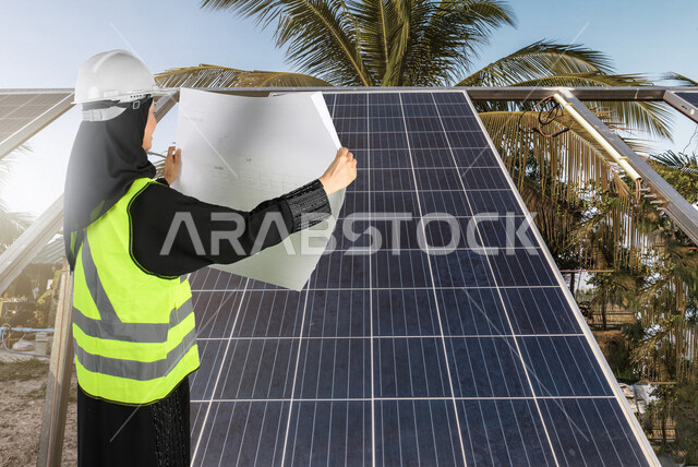صورة لمهندسة كهربائية عربية خليجية سعودية تحمل بيدها لوحة تصميم و مخطط خاص بالخلايا الشمسية ، تقف بجانب ألواح للخلايا الشمسية في مزرعة ، استخدام الطاقة الشمسية لتوليد الكهرباء ، مفهوم استخدام الطاقة المتجددة ، مفهوم التنمية المستدامة بالطاقة البديلة
