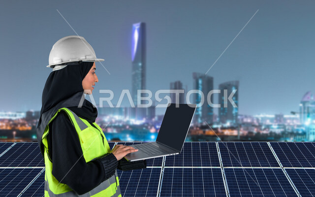 صورة لمهندسة كهربائية عربية خليجية سعودية تحمل حاسوب محمول في يدها تقف بالقرب من ألواح للخلايا الشمسية ، بخلفية لابراج المملكة العربية السعودية ، استخدام الطاقة الشمسية لتوليد الكهرباء ، مفهوم استخدام الطاقة المتجددة ، مفهوم التنمية المستدامة بالطاقة البد