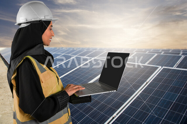 صورة لمهندسة كهربائية عربية خليجية سعودية تحمل حاسوب محمول في يدها تقف بالقرب من ألواح للخلايا الشمسية ، استخدام الطاقة الشمسية لتوليد الكهرباء ، مفهوم استخدام الطاقة المتجددة ، مفهوم التنمية المستدامة بالطاقة البديلة