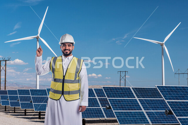 صورة لمهندس كهربائي عربي خليجي سعودي مبتسم ، يقف أمام لوحات للخلايا الشمسية لتوليد الكهرباء و طوحين الهواء ، يشير بيده نحوها ، ايماءات وتعابير تدل على السعادة ، مفهوم استخدام الطاقة المتجددة ، مفهوم التكنولوجيا التنمية المستدامة بالطاقة البديلة