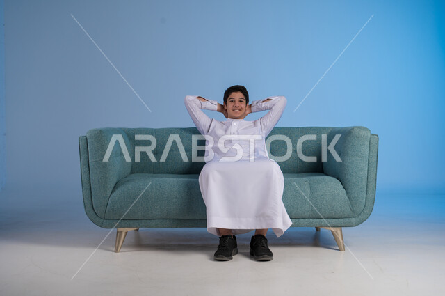 مفهوم الراحة والاسترخاء, الاستمتاع بالأجواء الهادئة, الشعور بالتعب والخمول, بورتريه لشاب عربي خليجي سعودي يرتدي الثوب التقليدي يجلس على اريكة مريحة, خلفية زرقاء