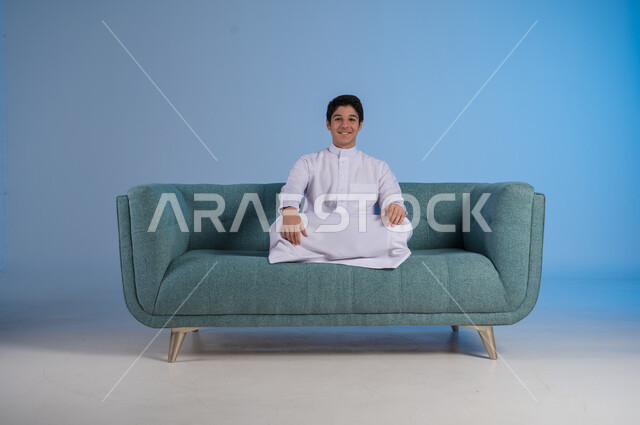 الاستمتاع بالأجواء الهادئة, مفهوم الراحة والاسترخاء, الشعور بالتعب والخمول, بورتريه لشاب عربي خليجي سعودي يرتدي الثوب التقليدي يجلس على اريكة مريحة, خلفية زرقاء