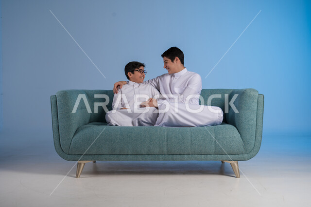 قضاء أوقات ممتعة مع الأخوات, تعزيز وتقوية العلاقة الإيجابية الأسرية, مفهوم الحب والحنان بين الأخوة, بورتريه لأخيين عربيين خليجين سعوديين يرتديان الثوب التقليدي يجلسان على كنبه مريحة, خلفية زرقاء