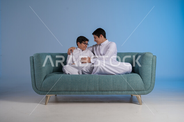 قضاء أوقات ممتعة مع الأخوات, تعزيز وتقوية العلاقة الإيجابية الأسرية, مفهوم الحب والحنان بين الأخوة, بورتريه لأخيين عربيين خليجين سعوديين يرتديان الثوب التقليدي يجلسان على كنبه مريحة, خلفية زرقاء