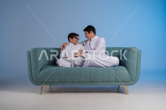 قضاء أوقات ممتعة مع الأخوات, تعزيز وتقوية العلاقة الإيجابية الأسرية, مفهوم الحب والحنان بين الأخوة, بورتريه لأخيين عربيين خليجين سعوديين يرتديان الثوب التقليدي يجلسان على كنبه مريحة, خلفية زرقاء