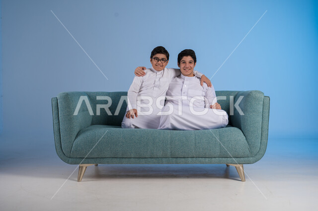النظر إلى الكاميرا بإيماءات السرور, أجواء ولحظات أسرية مليئة بالحب والحنان, مفهوم العائلة السعيدة, بورتريه لأخيين عربيين خليجين سعوديين يرتديان الثوب التقليدي يجلسان على كنبة مريحة, قضاء الوقت الممتع برفقة الأخوة, تعزيز العلاقات الأسرية الناجحة, خلفية زرقاء