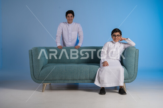 قضاء الوقت الممتع برفقة الأخوات, مفهوم تعزيز وتقوية الروابط الأسرية الناجحة, بورتريه لأخيين عربيين خليجيين سعوديين يرتديان الثوب التقليدي يستمتعان بلحظات عائلية لطيفة, خلفية زرقاء