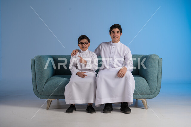 النظر إلى الكاميرا بإيماءات السرور, بورتريه لفتى عربي خليجي سعودي يرتدي الثوب التقليدي يشير إلى شيء ما, الجلوس في غرفة المعيشة والاستمتاع بالجلسات الأسرية مع الأخوات, خلفية زرقاء