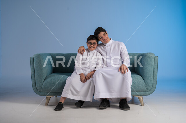 التسلية وقضاء الوقت الممتع مع الأخوات, تقوية الروابط الاسرية, بورتريه لشاب عربي خليجي سعودي يرتدي الثوب التقليدي يضع يده على كتف أخيه, لحظات مليئة بالحنان والحب, الجلوس والنظر إلى الكاميرا, خلفية زرقاء